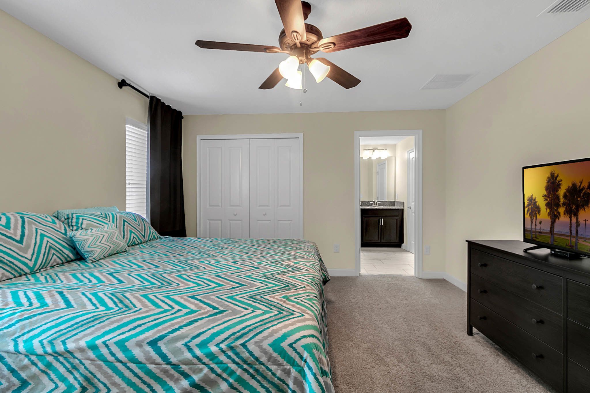 A bedroom with a bed, a ceiling fan, a dresser, and a door leading to a bathroom.