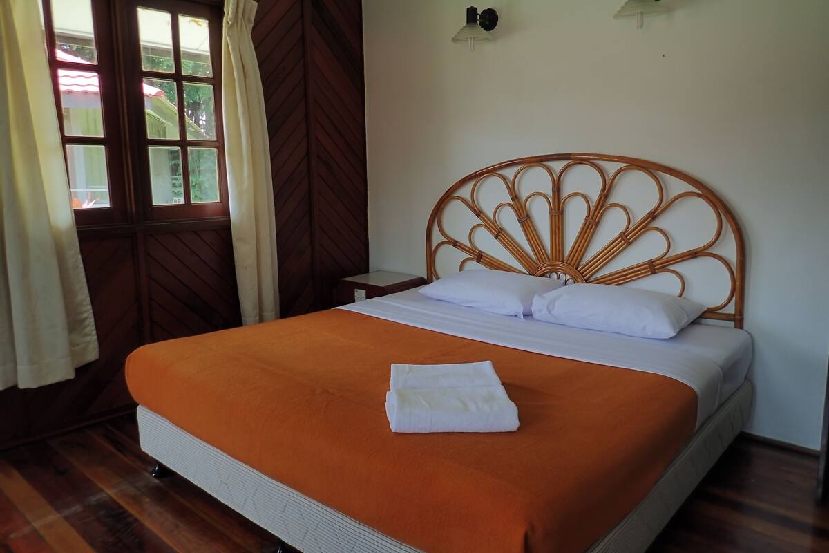 A neatly made bed with an orange bedspread and white pillows, a wooden headboard, a nightstand, and a window with curtains.