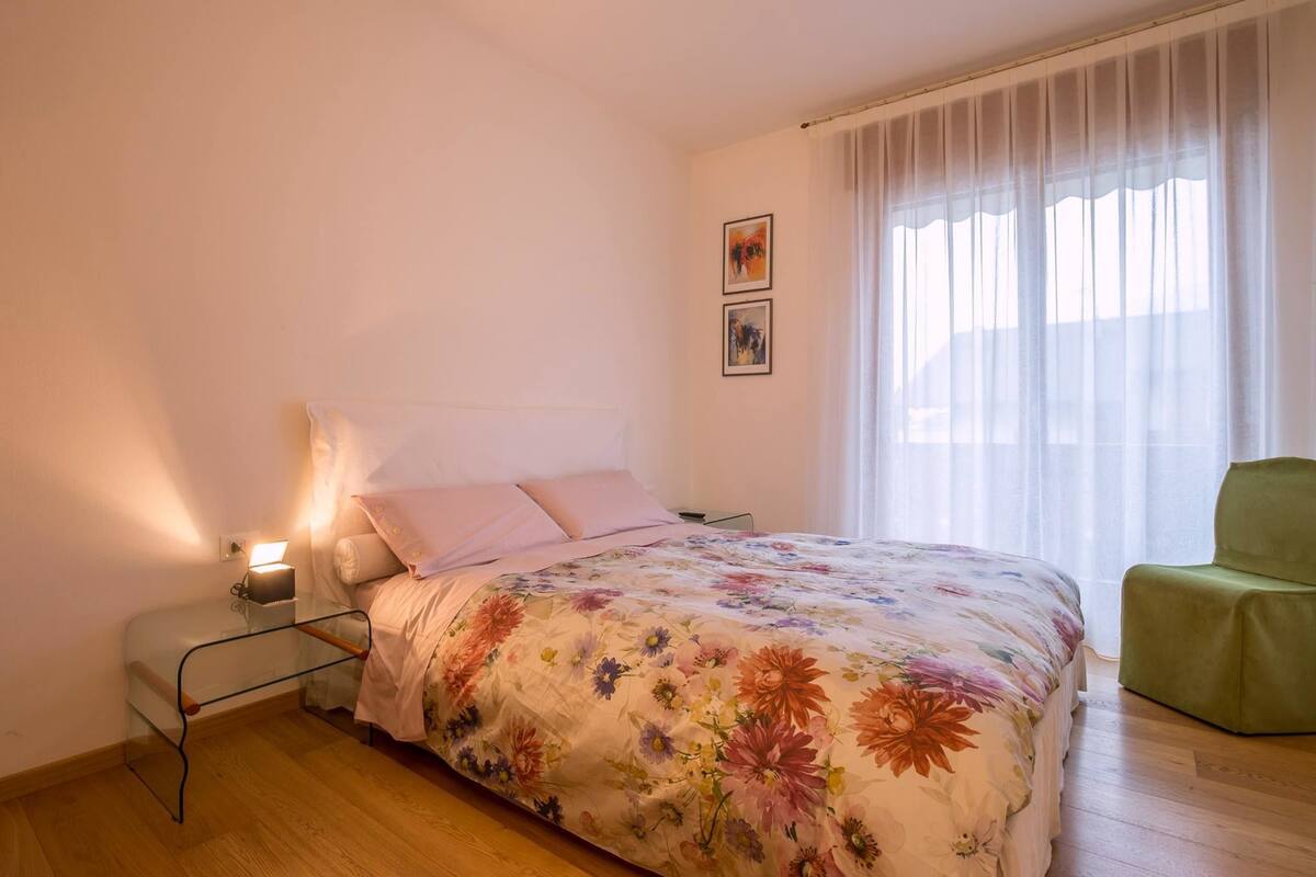 A bedroom with a floral bedspread, a bedside lamp, a green chair, and two framed pictures on the wall.