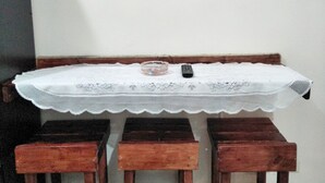Uma pequena área de jantar com uma mesa de madeira e três banquetas combinando, uma TV fixada na parede e uma toalha de mesa de renda.