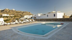 A swimming pool with lounge chairs, a stone wall, and white buildings in the background. - Evagelia's Place (Mykonos)