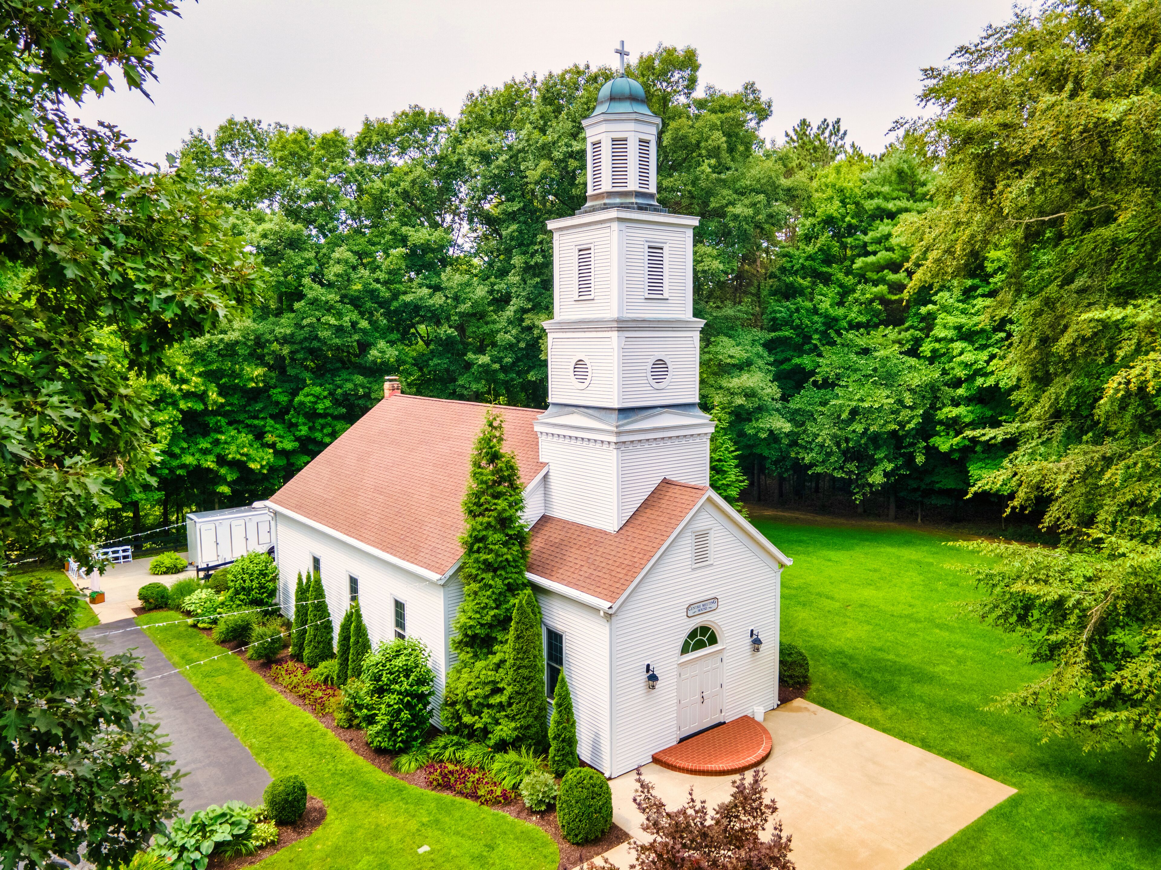 A white church with a bell tower and a red roof, surrounded by lush greenery.