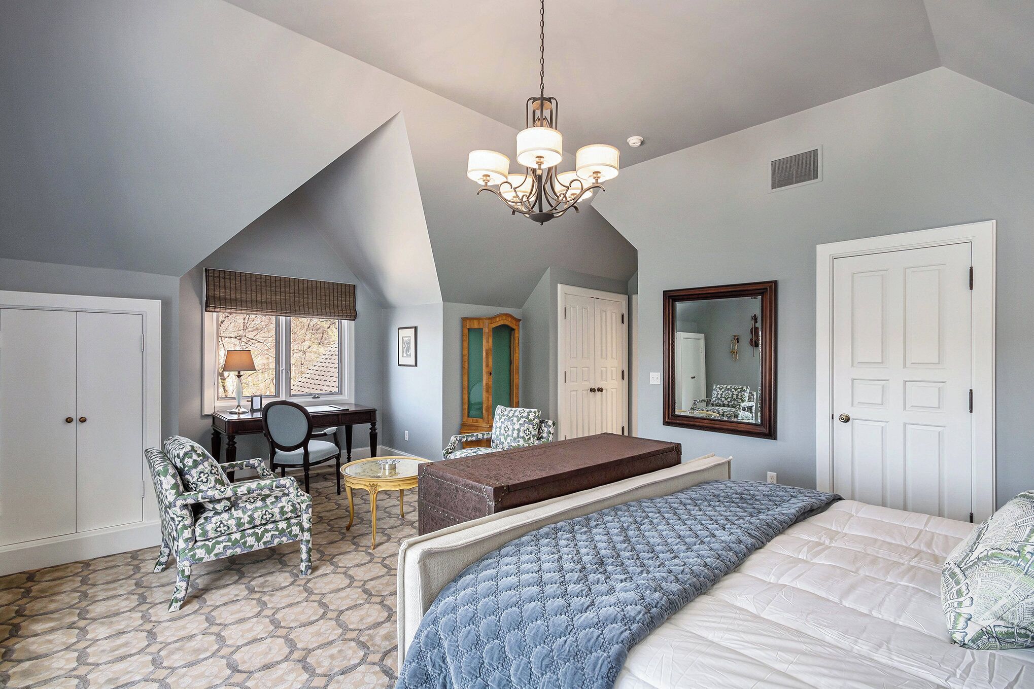 A bedroom with a bed, a desk, a chair, a small table, and a mirror.
