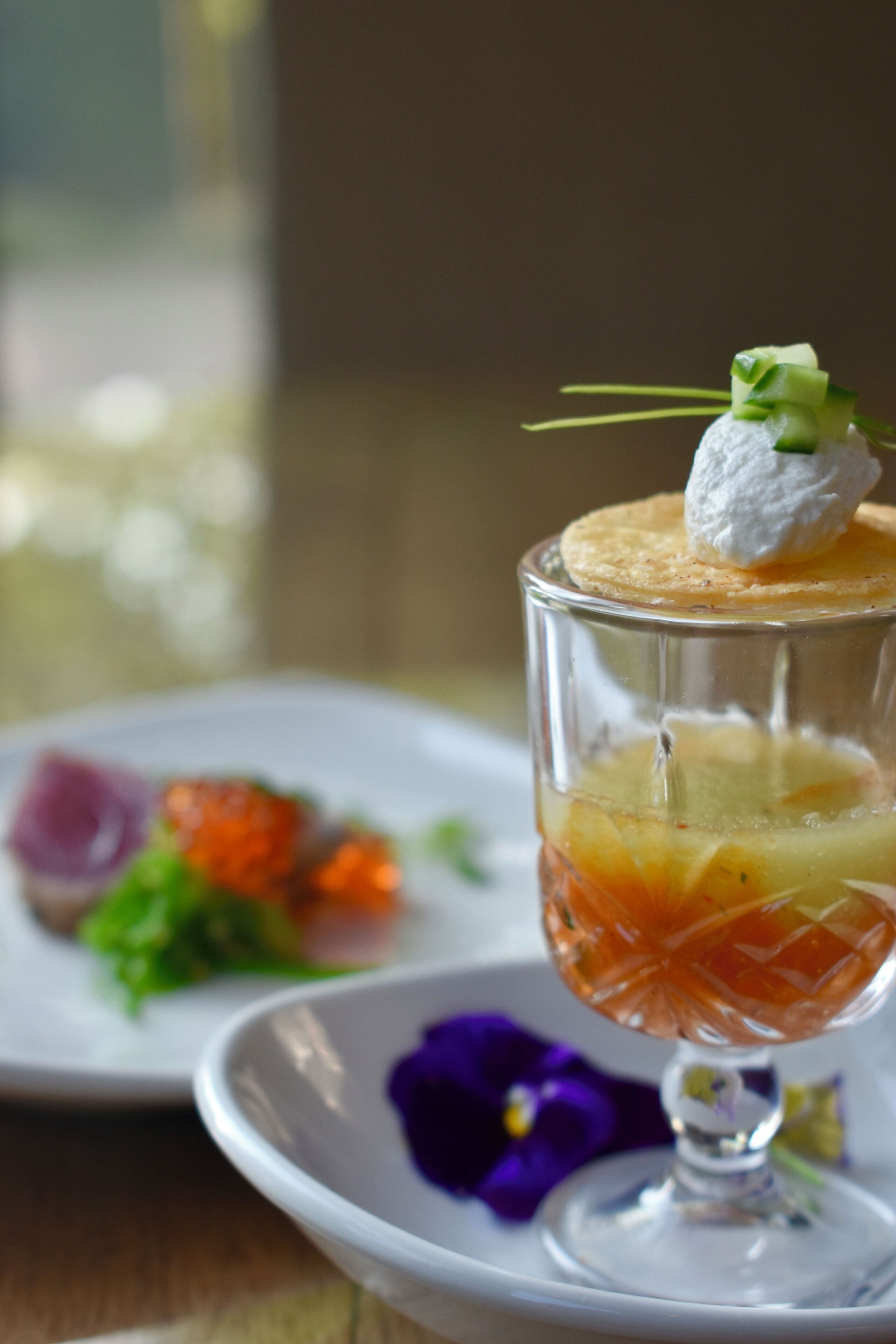 A dessert in a glass with a garnish on top, placed on a white plate with a purple flower beside it.