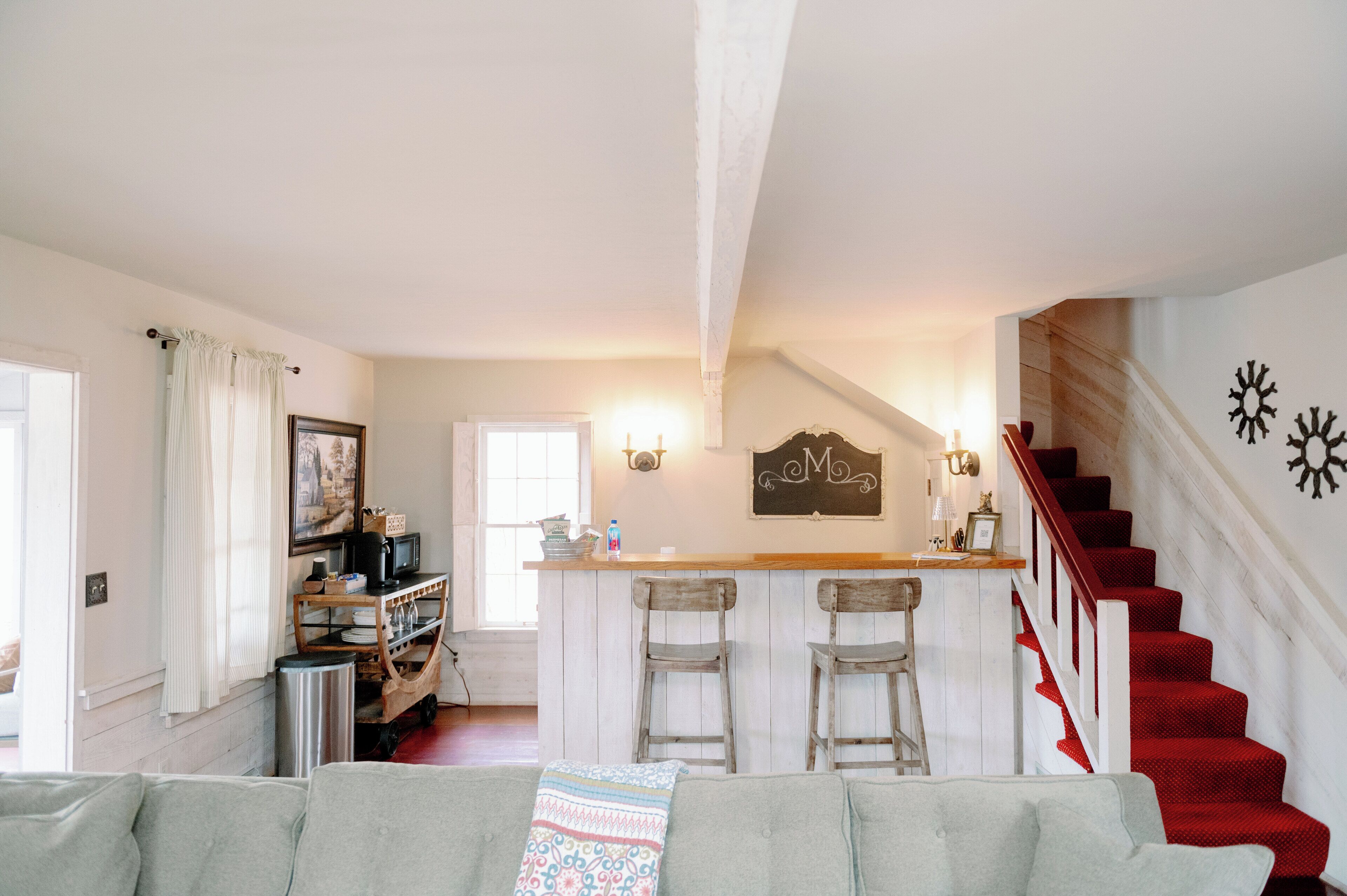 A cozy living room with a grey sofa, a wooden bar area with two stools, and a staircase with red carpeted steps.
