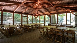 A covered outdoor dining area with wooden tables and chairs, string lights, and large windows offering views of the surrounding forest. - The Morris Estate (Niles)