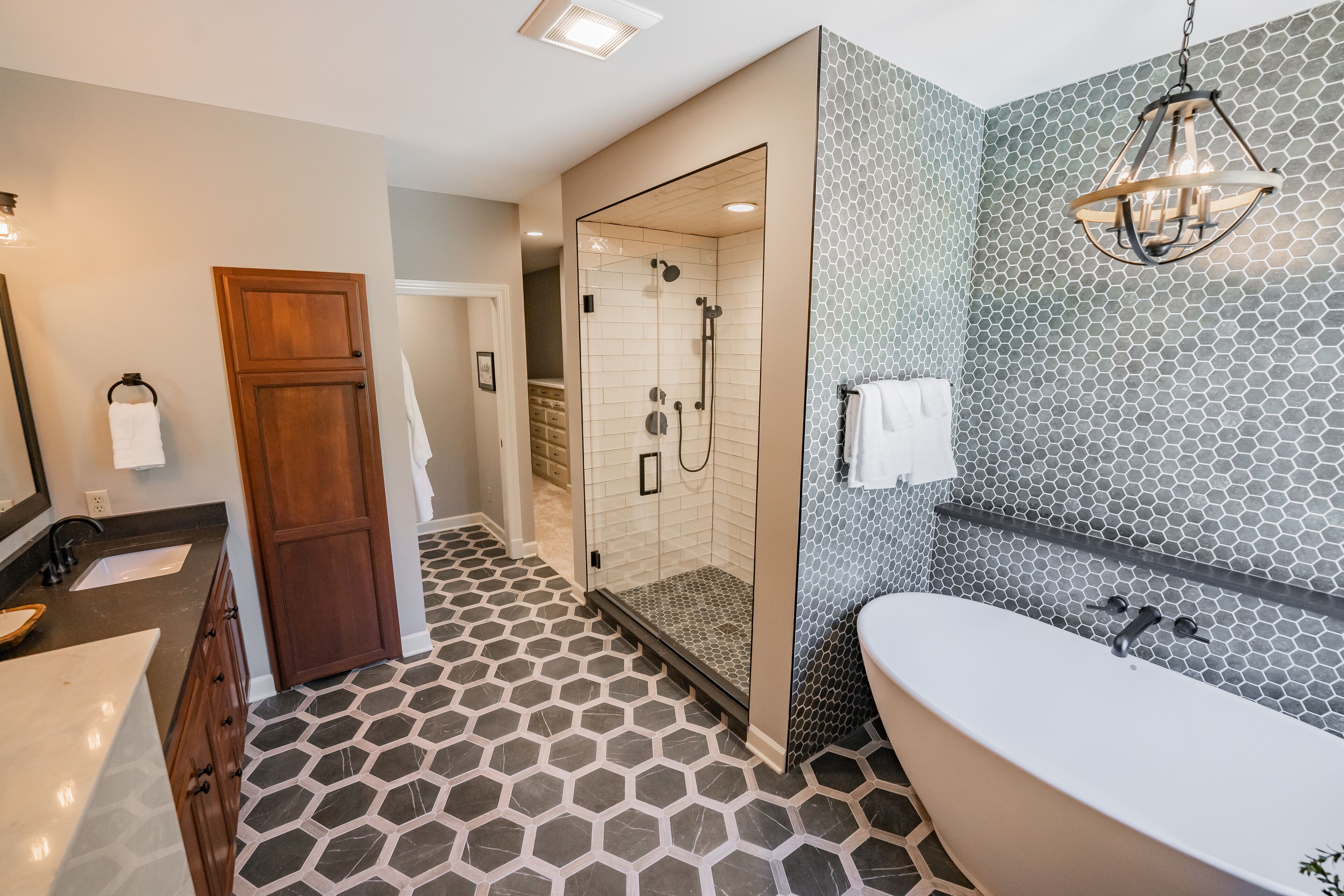 A modern bathroom with a hexagonal tile floor, a freestanding bathtub, a glass-enclosed shower, and a wooden door.