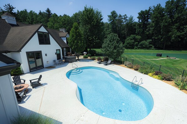 A residential swimming pool with a house, patio furniture, and a well-maintained lawn.
