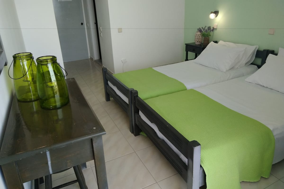Una habitación con una cama, una mesita de noche, una mesa con dos botellas de vidrio verde y una puerta.