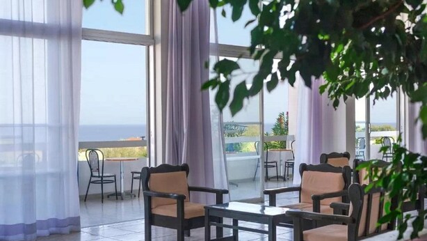 A terrace with chairs and a table, large windows with curtains, and a view of the sea. - Notos Hotel (Rhodes)