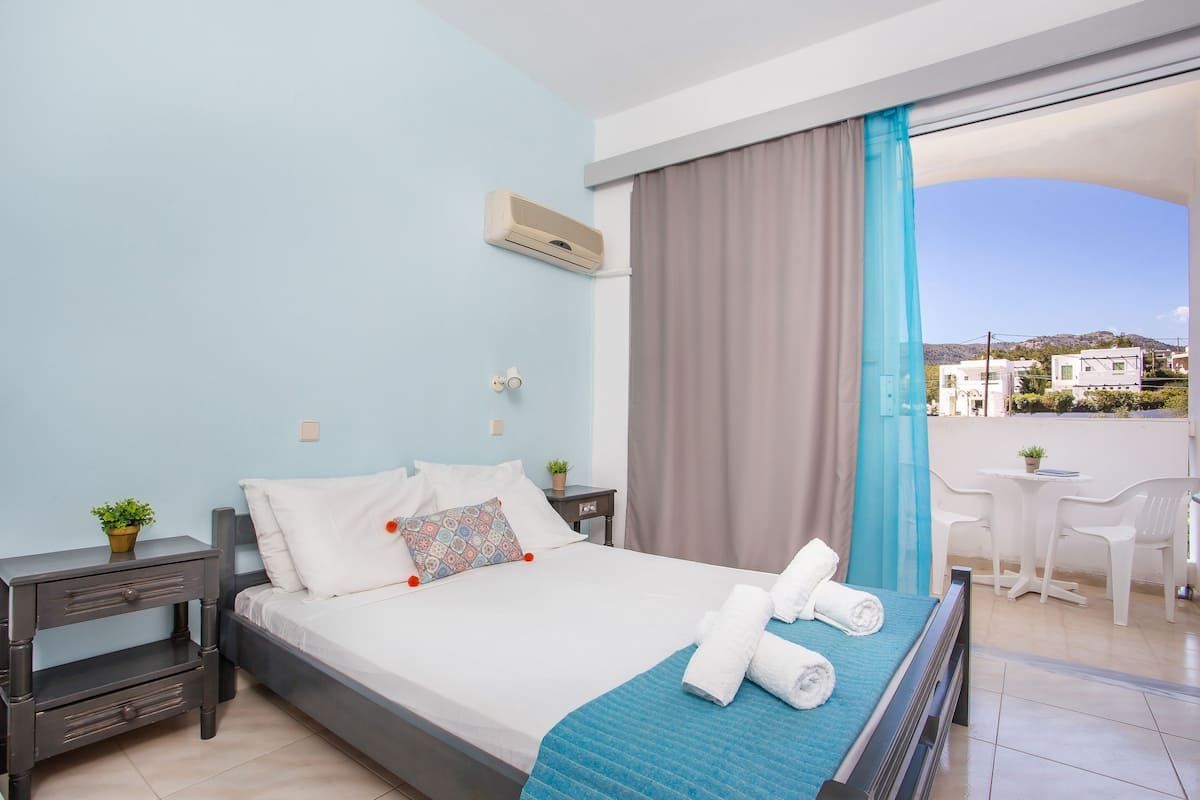 Habitación de hotel con cama, mesita de noche, planta en maceta, ventana con cortinas y balcón con sillas y mesa.