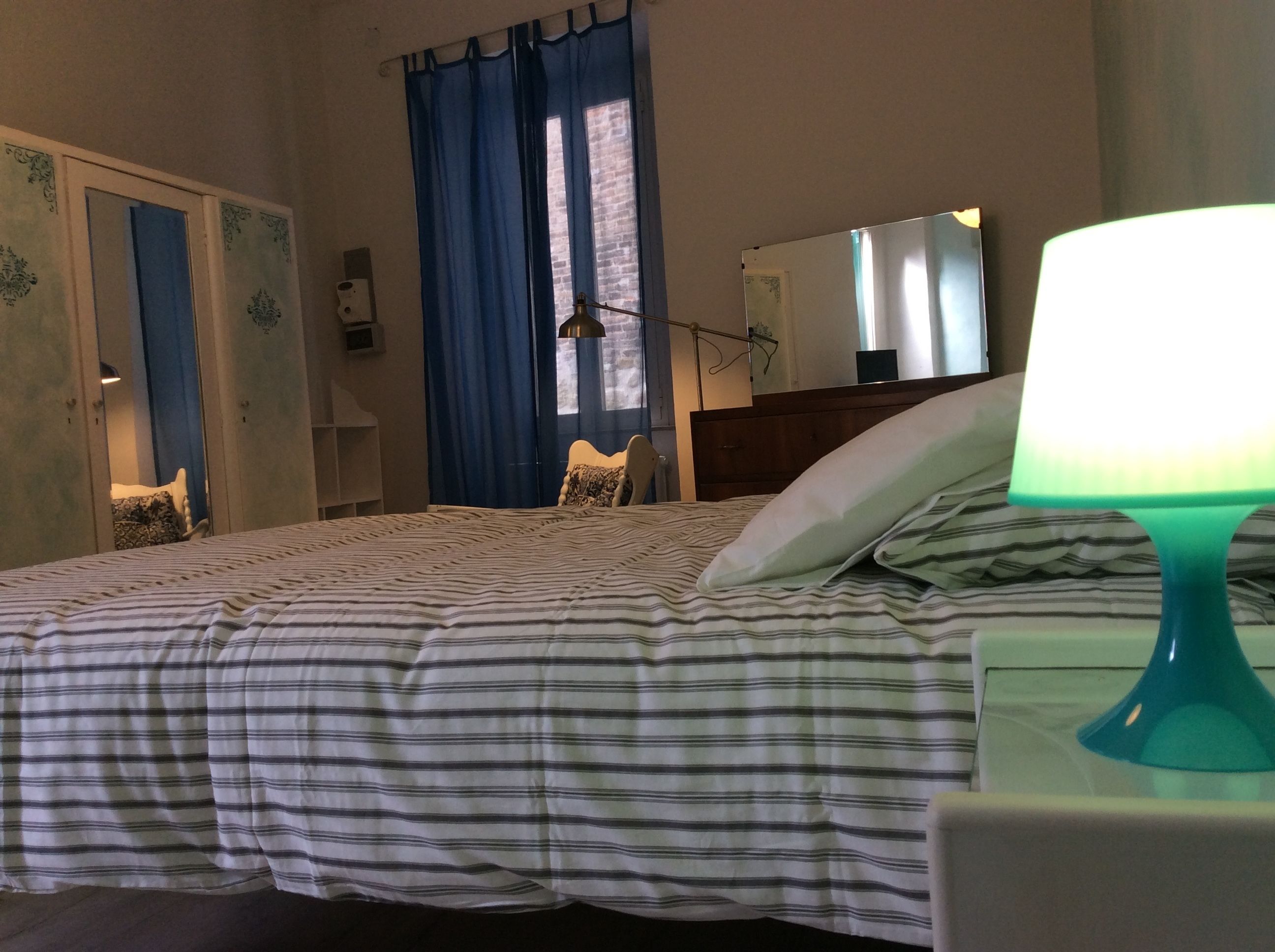 A bedroom with a bed, a nightstand, a lamp, a mirror, and a window with curtains.