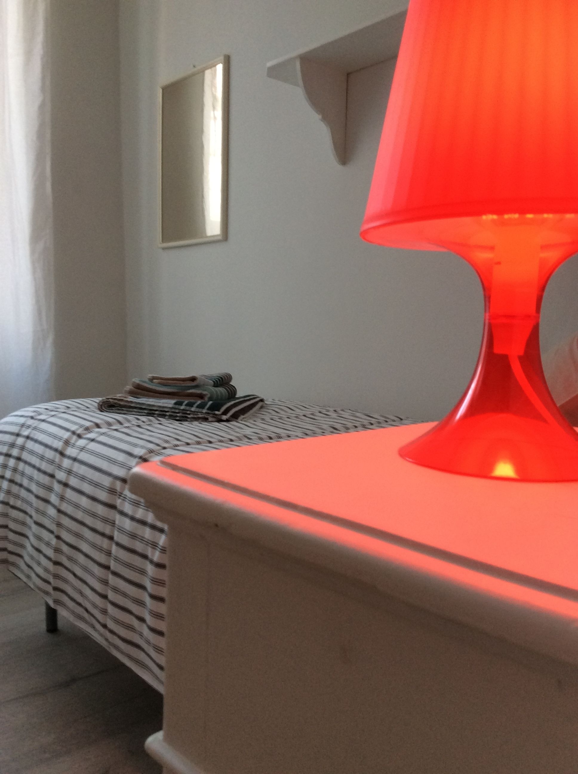 En seng med et stribet sengetæppe, et natbord med en rød lampe og et spejl på væggen.