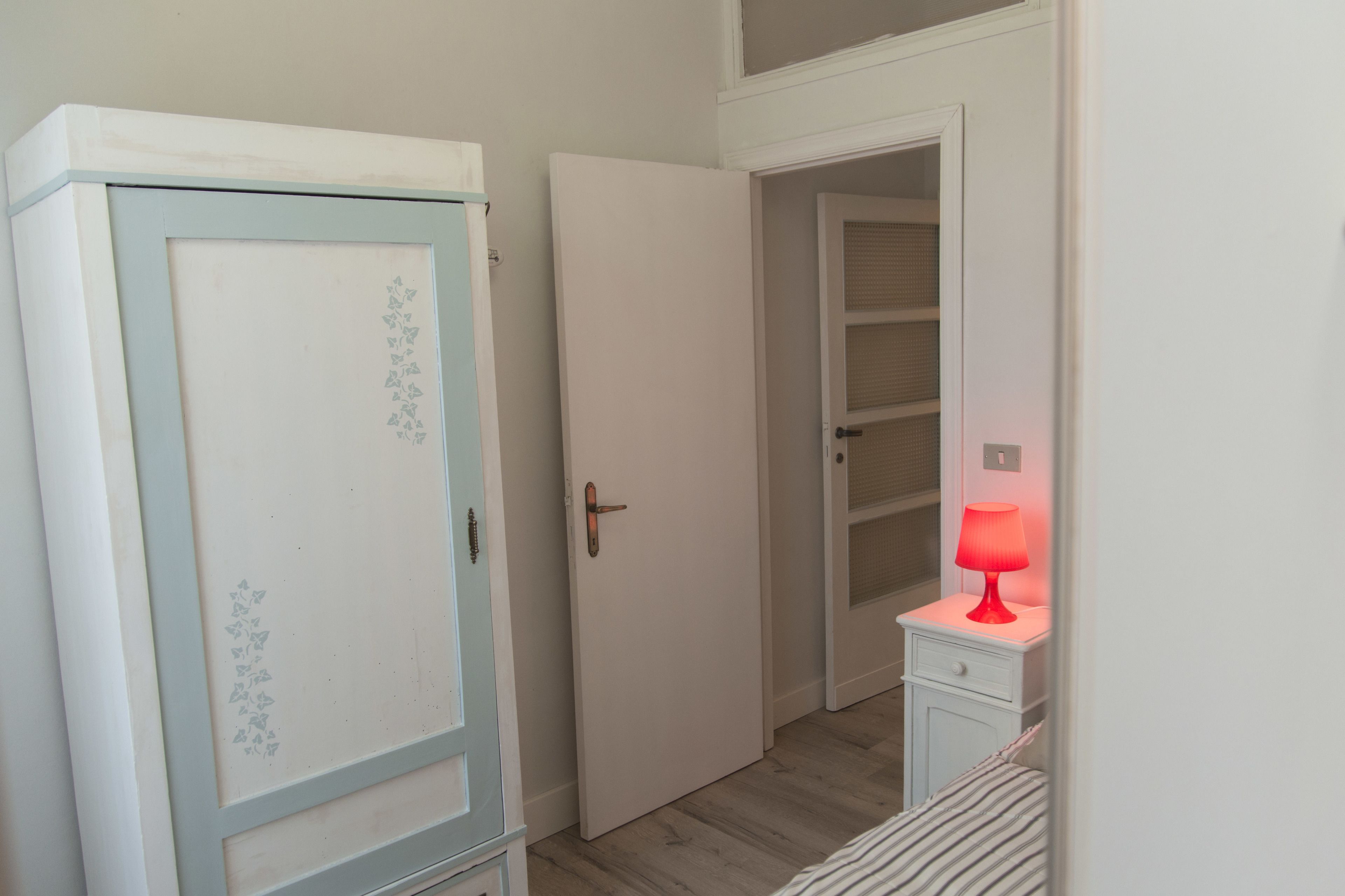 Een smalle gang met een witte deur, een garderobe met lichtblauwe accenten en een nachtkastje met een rode lamp.
