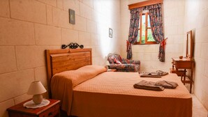 A bedroom with a wooden bed, a nightstand with a lamp, a chair, a small table, and a window with curtains. - Country View (Ghasri)