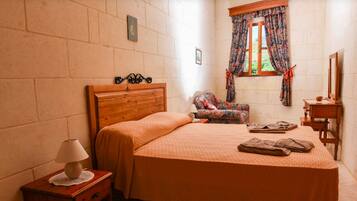A bedroom with a wooden bed, a nightstand with a lamp, a chair, a small table, and a window with curtains.