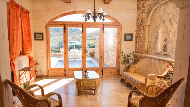 A room with a wooden table, wicker chairs, a sofa, and a view of a pool and mountains through large glass doors. - Country View (Ghasri)