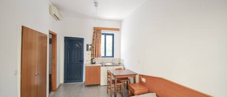 A compact room with a bed, a small table, a chair, a kitchenette, and a door leading to another room.