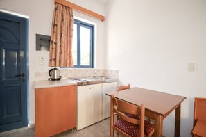 Eine kleine Küche mit Spüle, Herd und Schränken. Es gibt ein Fenster mit Vorhängen, eine Tür und einen kleinen Tisch mit Stühlen.