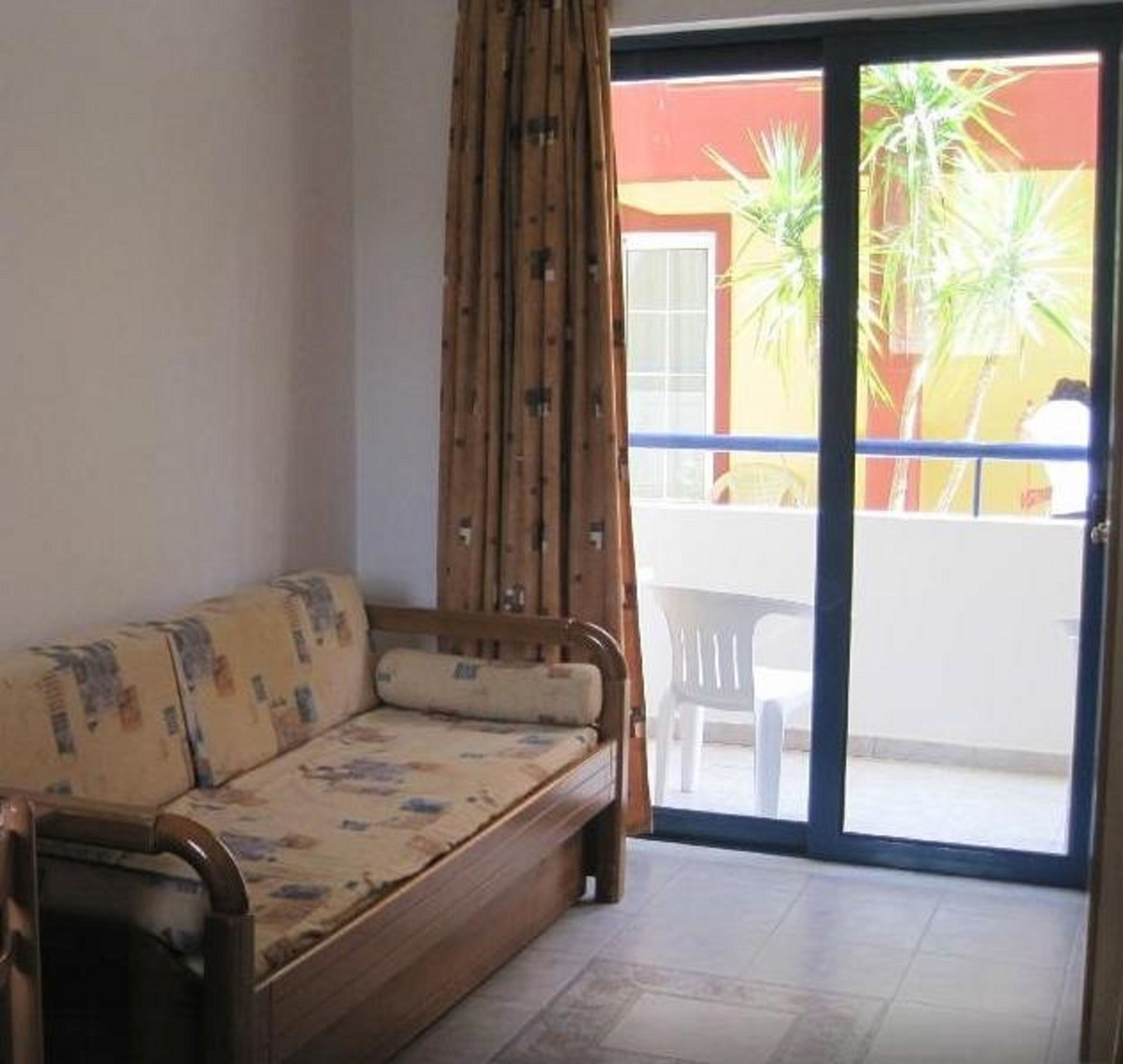 Una habitación con sofá de madera, una cortina y un ventanal corredero que da a un espacio exterior con una mesa y sillas.