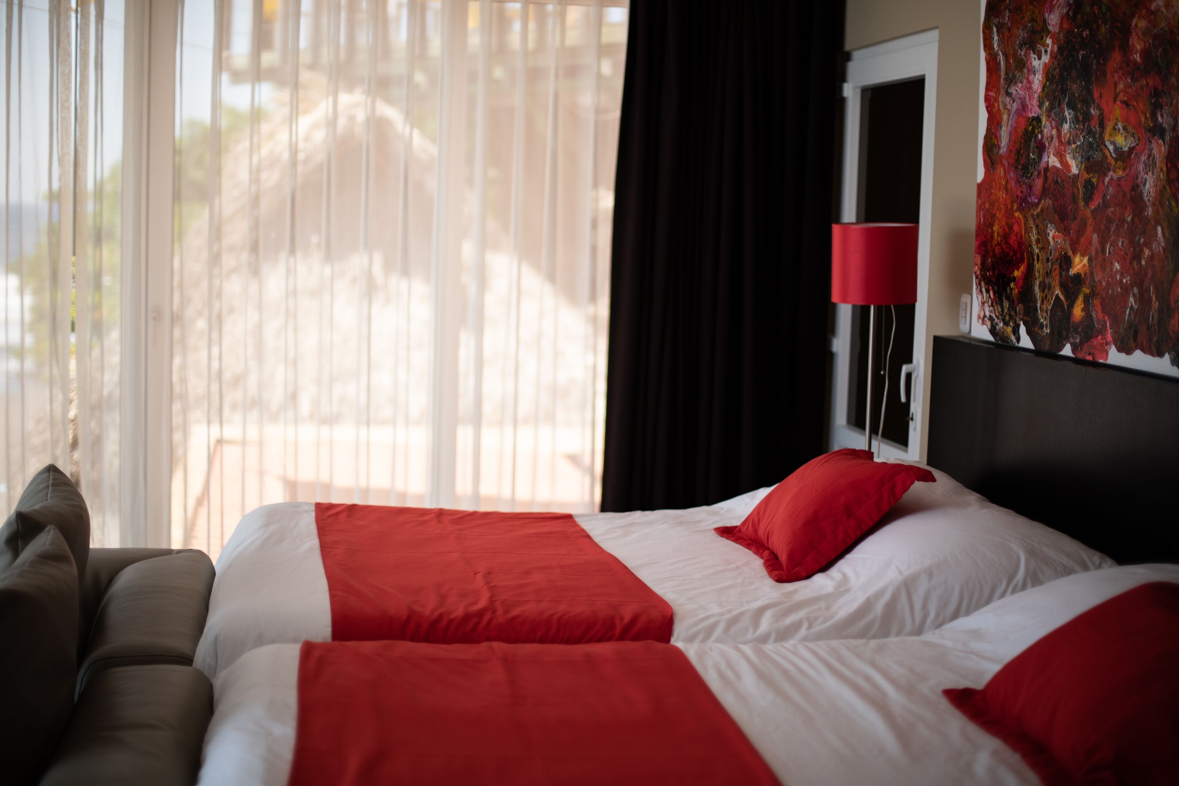 A hotel room with a bed, a red lamp, and a large abstract painting on the wall.