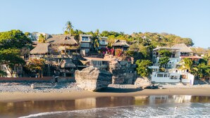 A beachfront property with a rocky shoreline, a person surfing, and buildings with balconies overlooking the beach. - Casa de Mar Hotel And Villas (Tamanique)