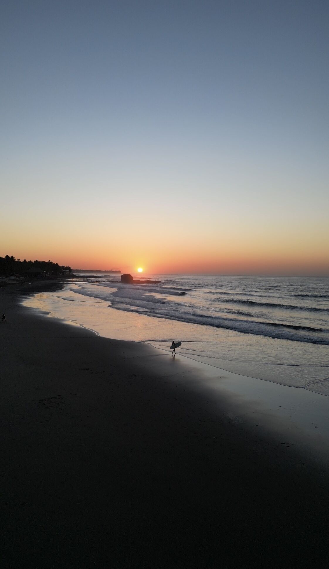 A beach at sunset with calm waves and a person with a surfboard.