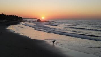 A beach at sunset with calm waves and a person with a surfboard.