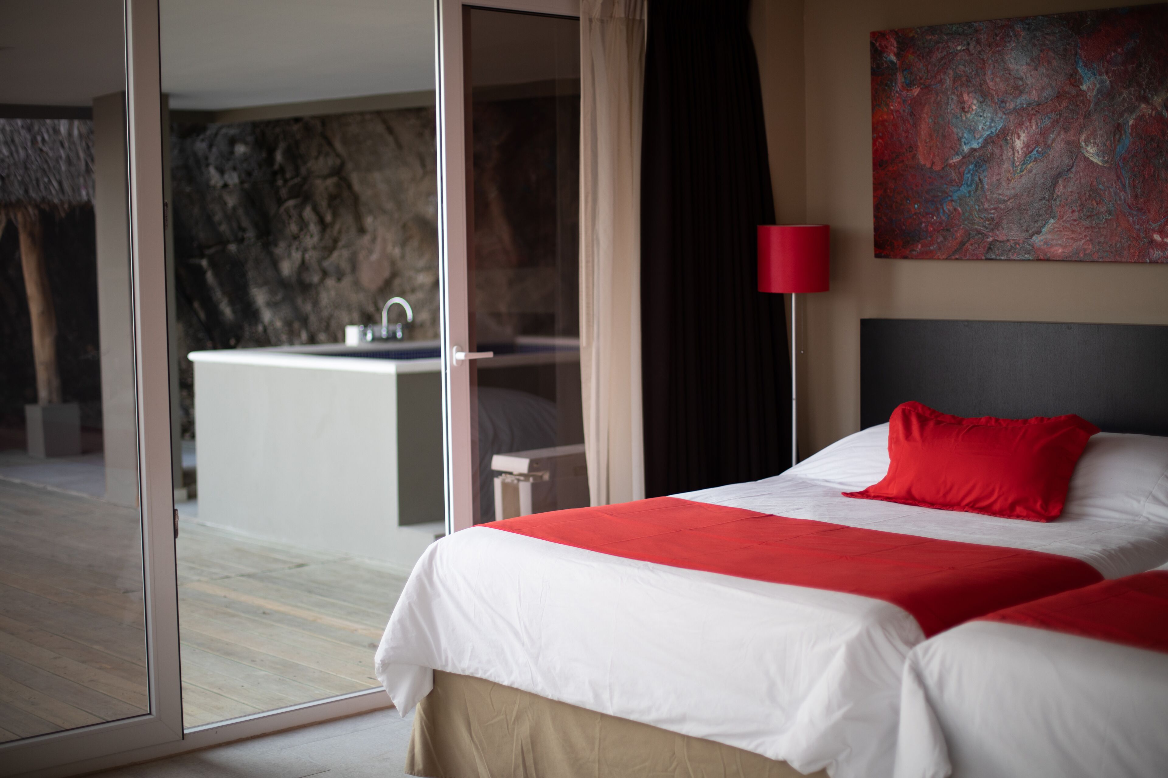 A hotel room with a large bed, a red pillow, a red lamp, and a view of a bathroom with a bathtub.