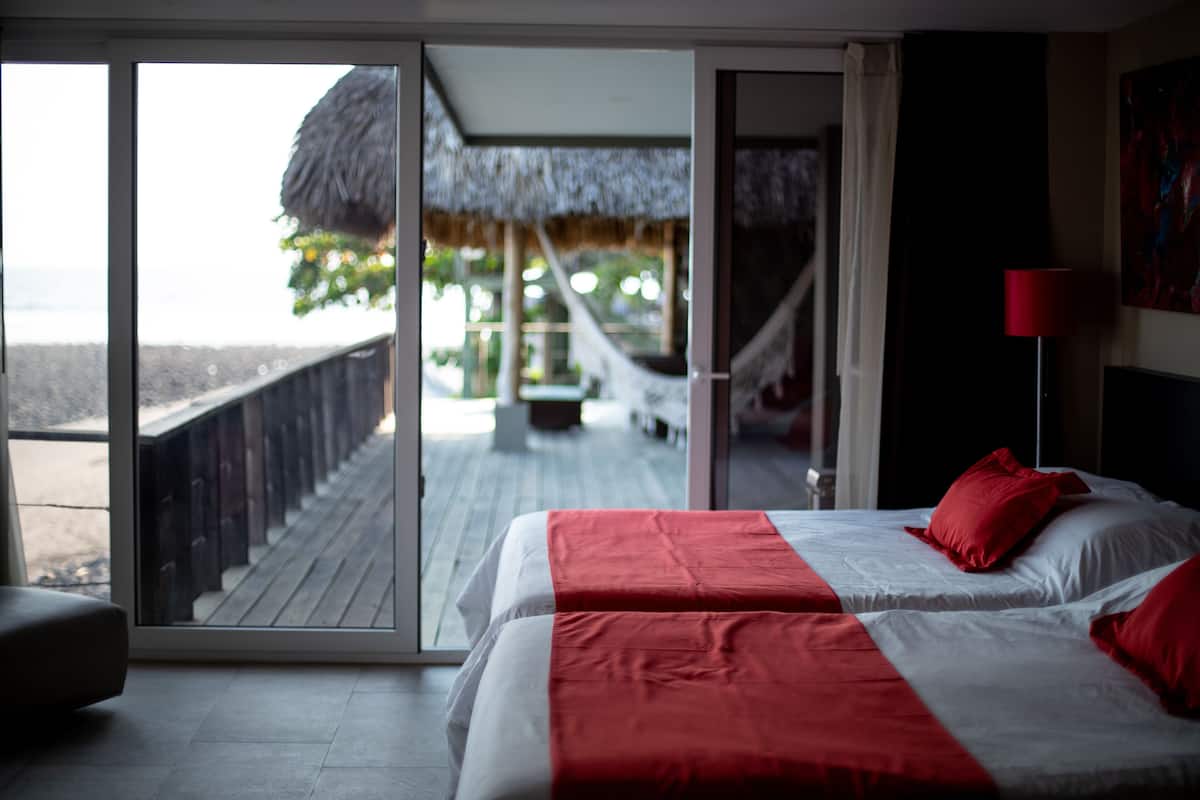 Une chambre d’hôtel avec un lit, une couvre-lit rouge et blanche, une lampe rouge et une vue sur une plage et une structure à toit de chaume, visible à travers une porte vitrée.