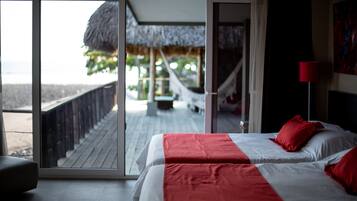 Quarto de hotel com uma cama, um edredom vermelho e branco, uma luminária vermelha e vista para uma praia e uma construção com telhado de palha através de uma porta de vidro.