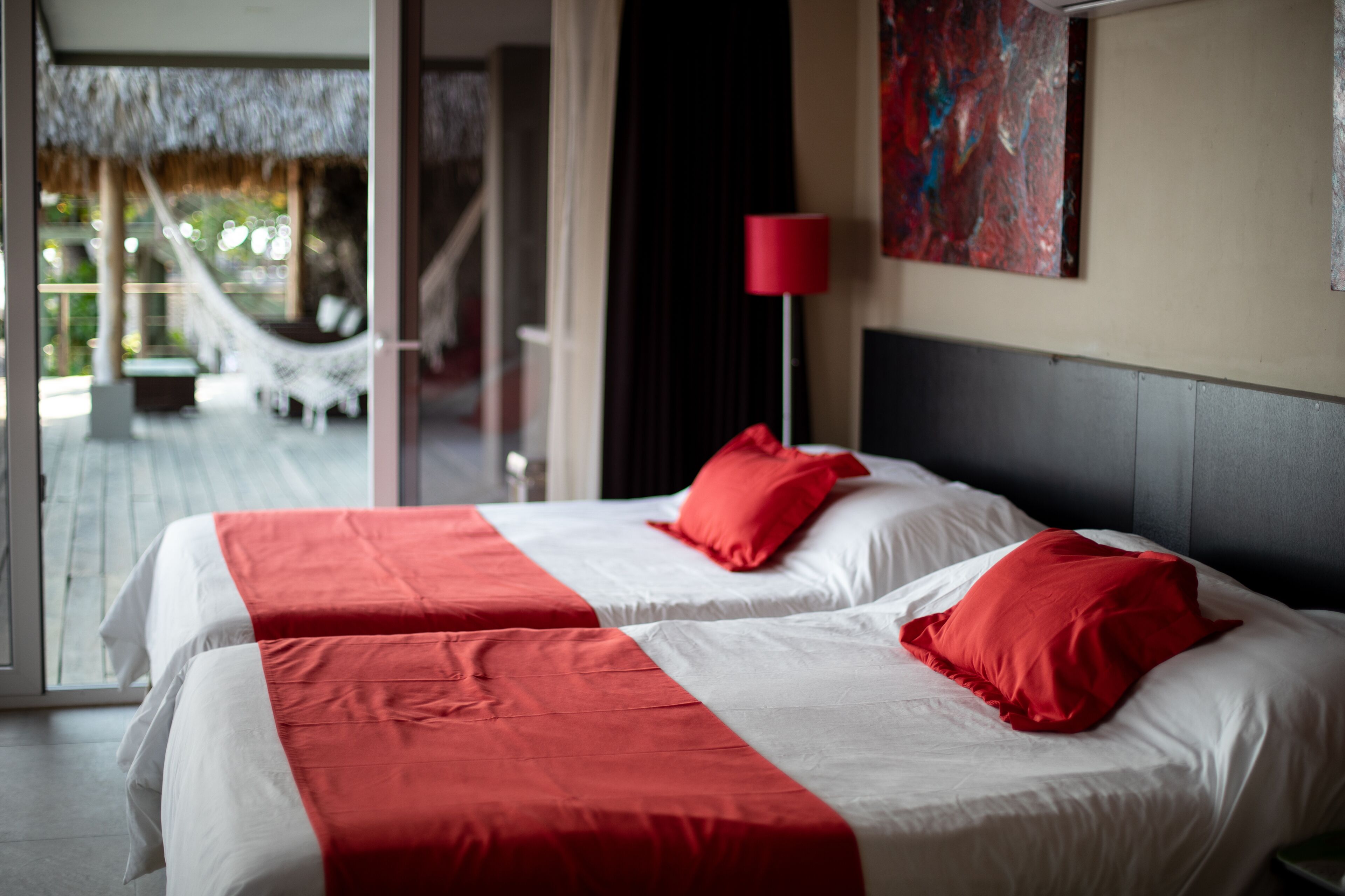 A hotel room with two beds, each with red and white bedding, a red floor lamp, and a painting on the wall.