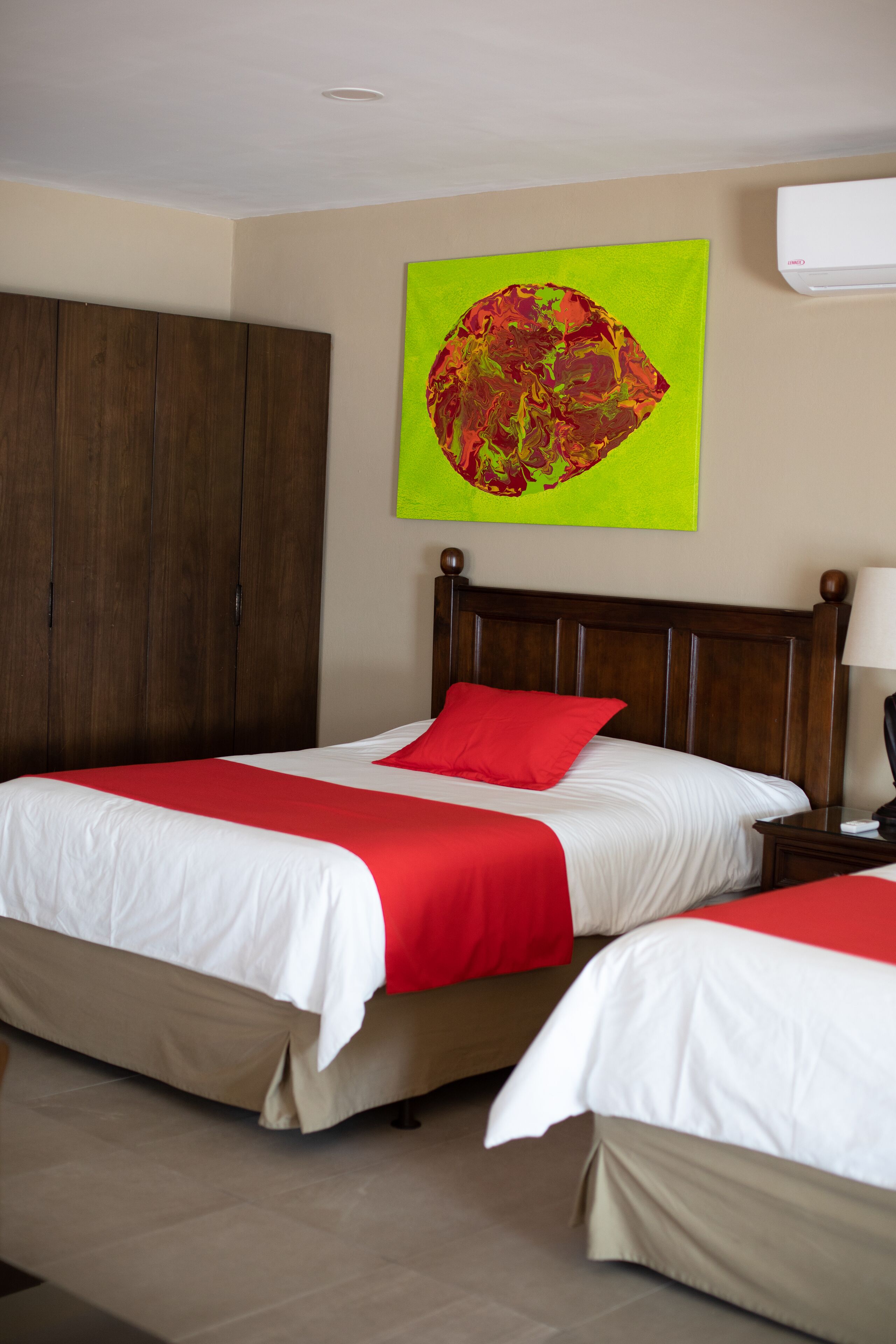 Habitación de hotel con dos camas, cabecero de madera, colcha roja y blanca, cuadro abstracto verde en la pared y unidad de aire acondicionado blanca.