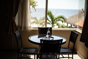 A round table with four chairs, a bottle, and a glass. The room has a large window with a view of the ocean and palm trees.
