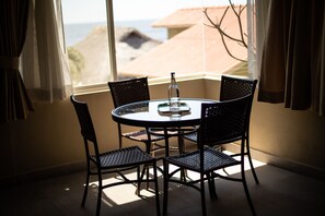 A round table with four chairs, a bottle, and a glass, positioned by a window with curtains, overlooking a beach and buildings.