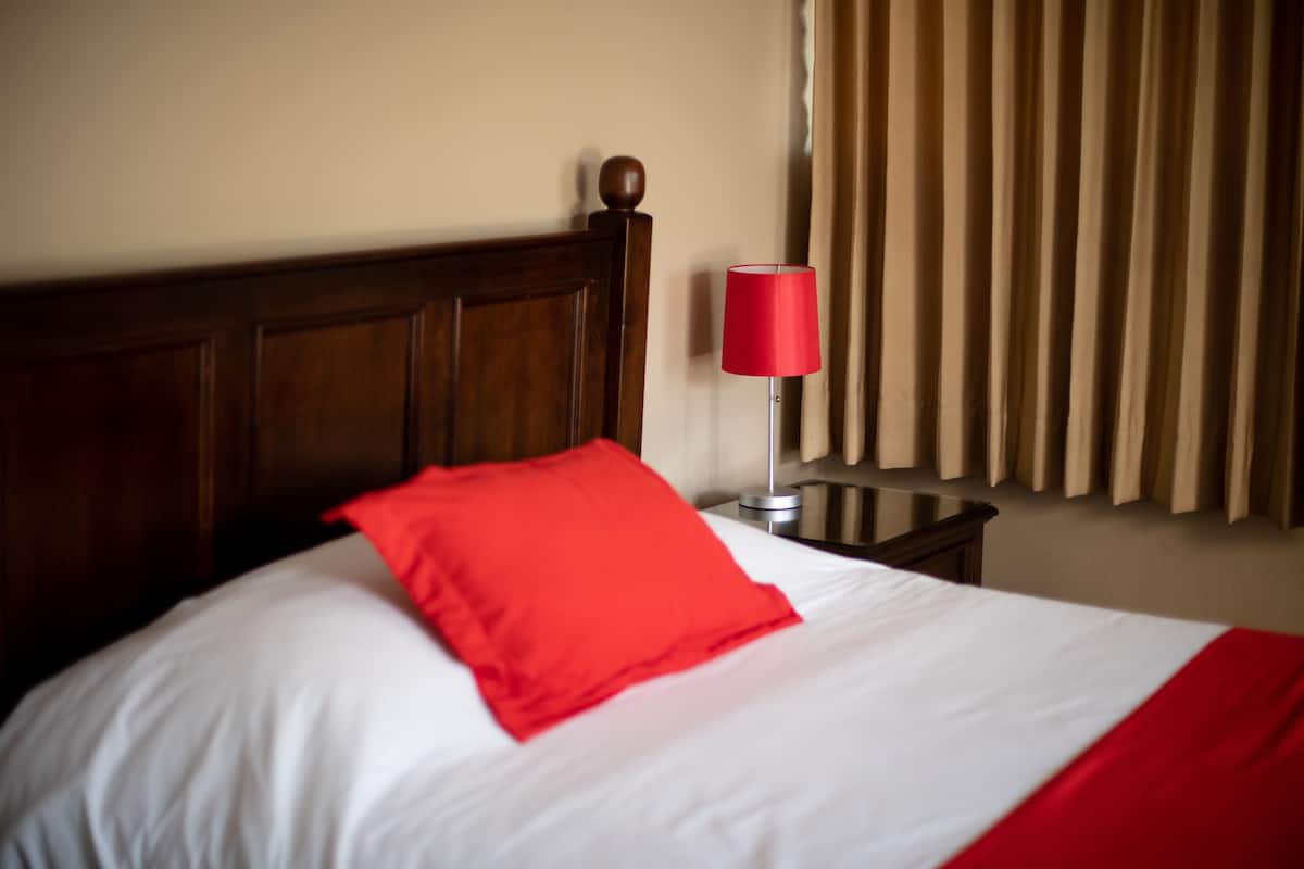 Une chambre d’hôtel avec une tête de lit en bois, un oreiller rouge, un couvre-lit blanc, une lampe rouge et une fenêtre avec des stores verticaux.