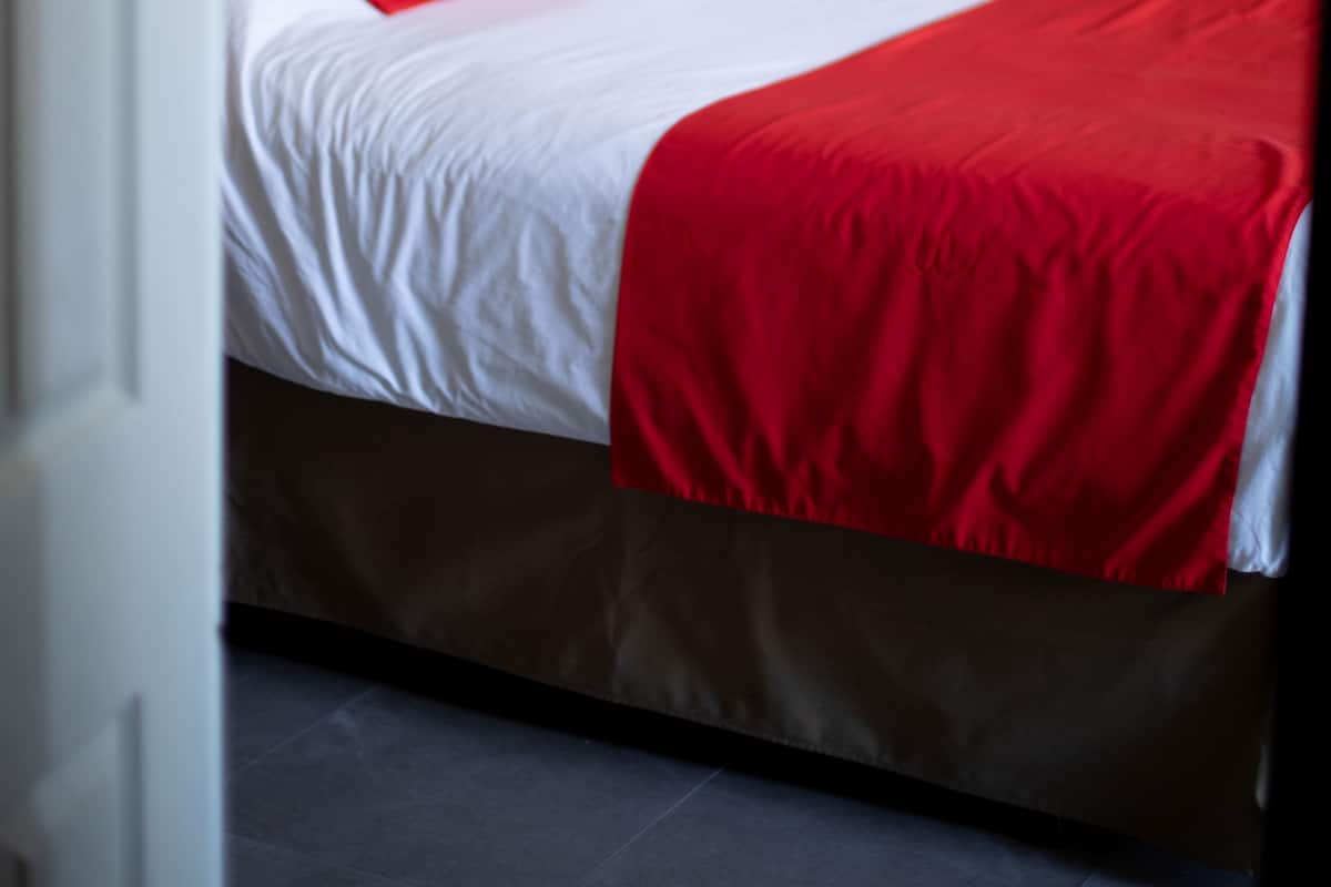 Un lit avec une couvre-lit blanche et rouge, une tête de lit en bois et une lampe rouge posée sur une table de chevet.