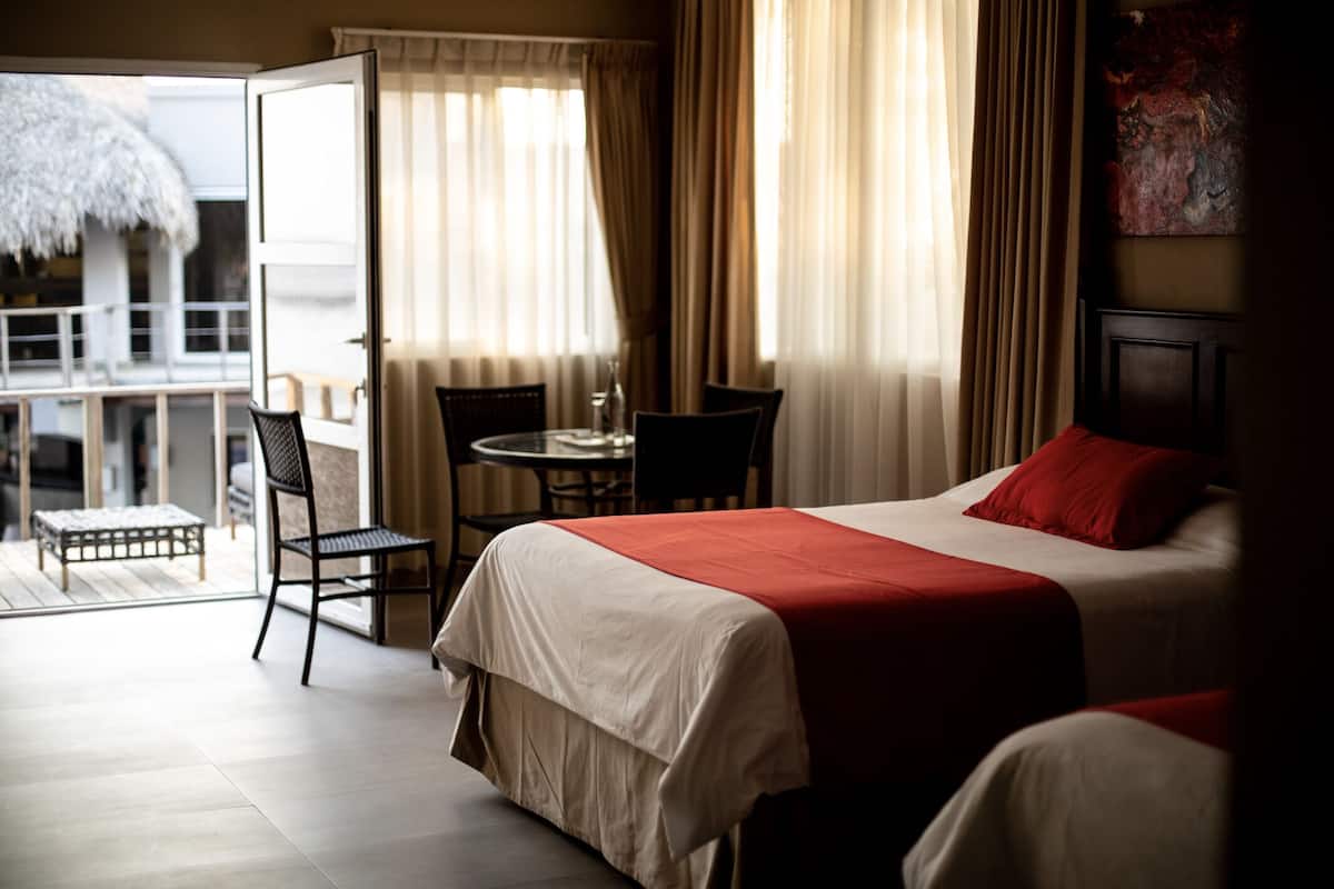 Une chambre d’hôtel avec un lit, une chaise, une petite table et un balcon agrémenté d’un parasol en chaume.