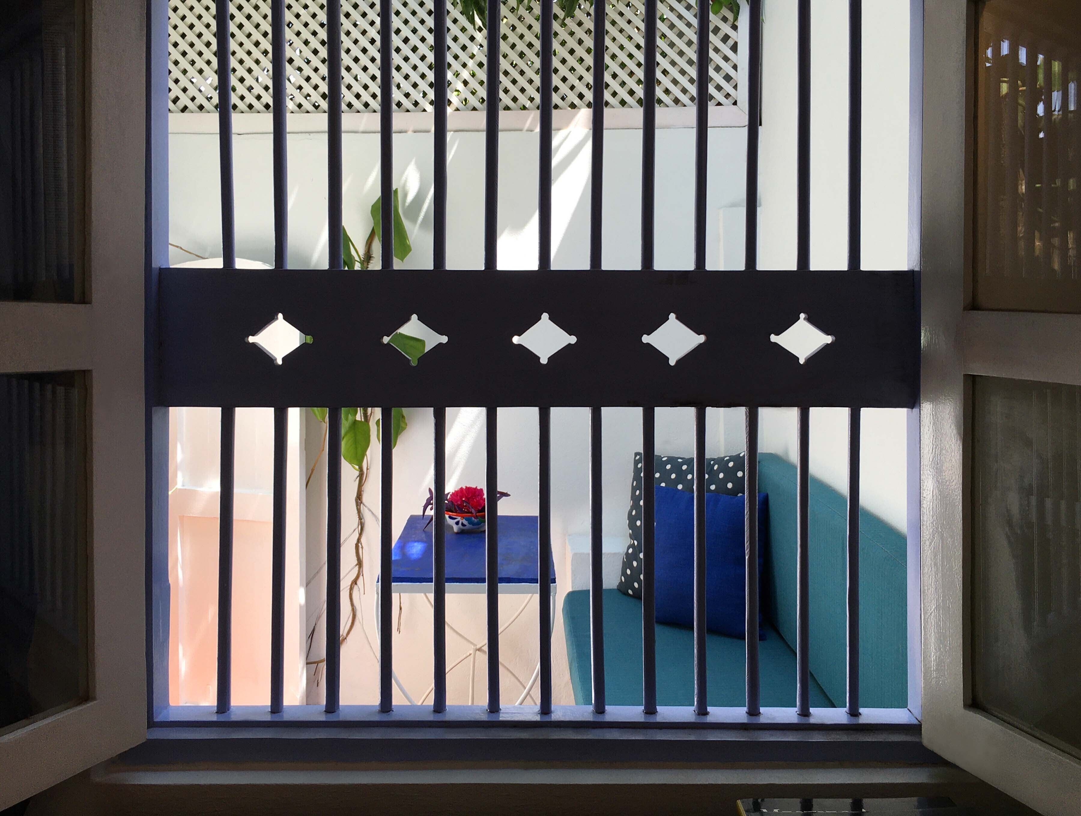 Une pièce dotée d’une fenêtre ornée d’une grille en métal avec des motifs décoratifs découpés, d’un canapé bleu agrémenté d’un coussin à pois et d’une petite table sur laquelle est posé un vase avec une fleur rouge.