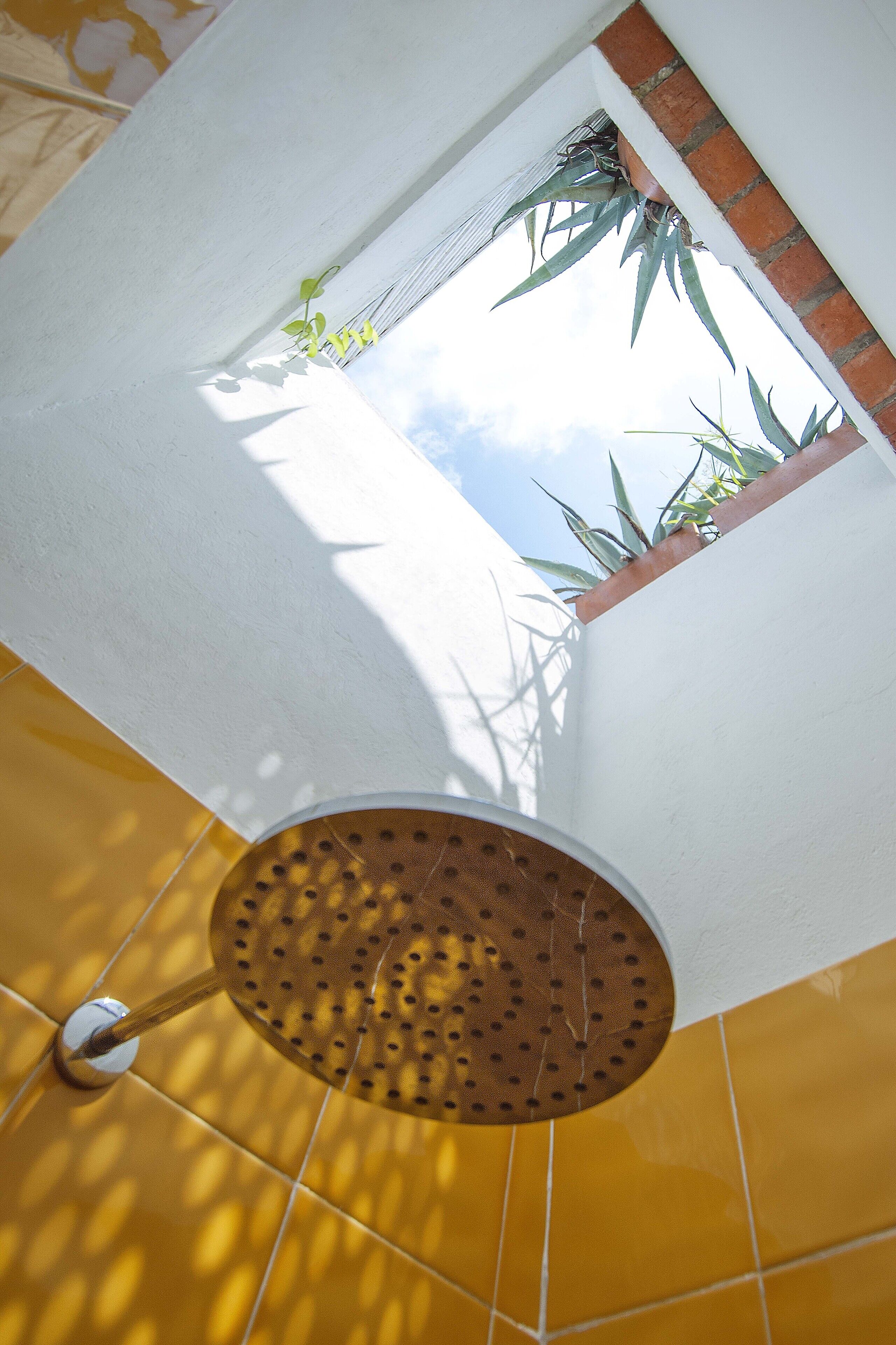 A corner of a room with a white wall, a yellow tiled floor, and a window with a view of the sky and plants outside.