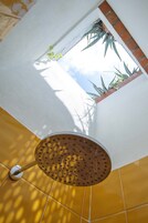A corner of a room with a white wall, a yellow tiled floor, and a window with a view of the sky and plants outside.