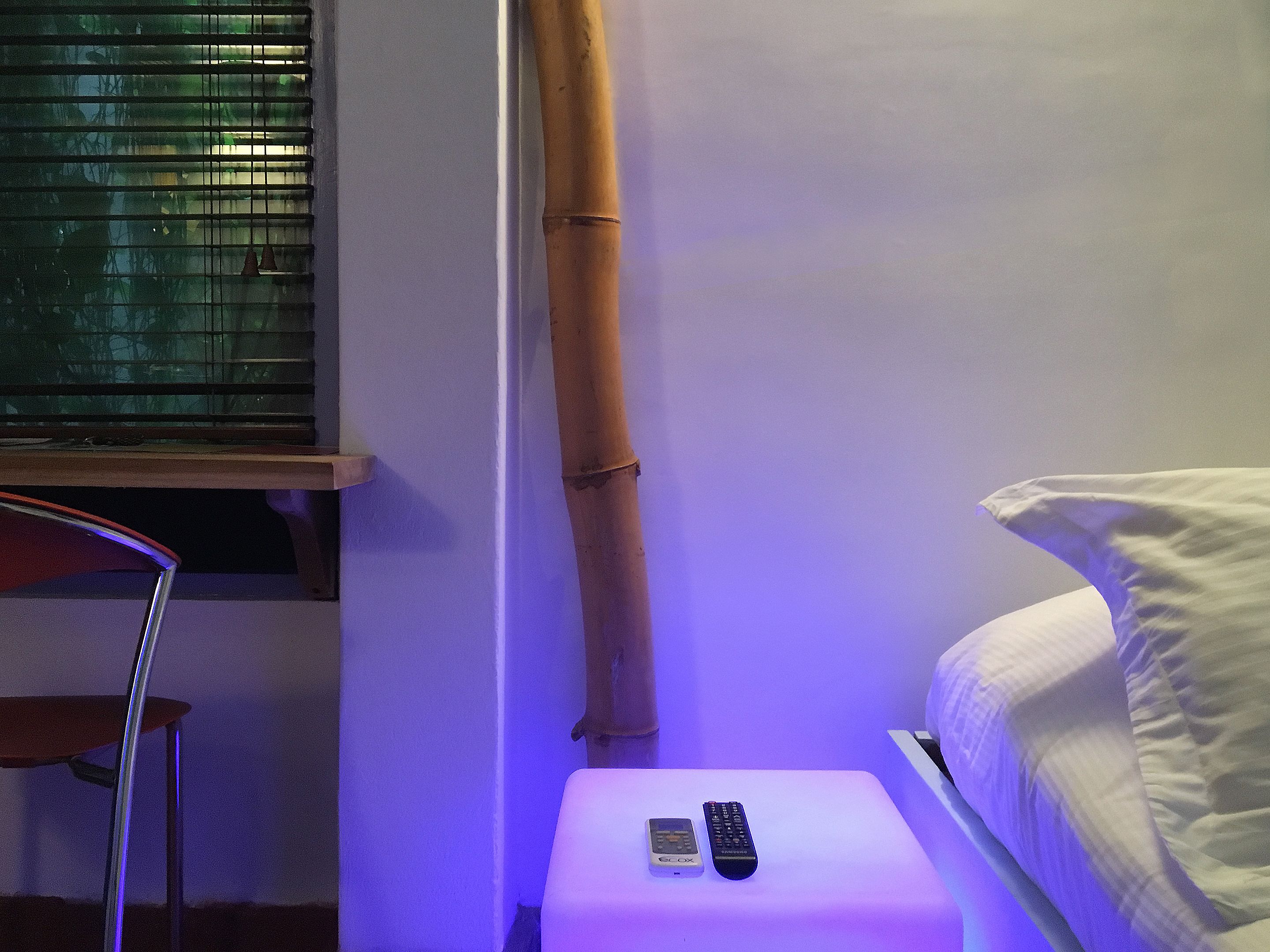 A room with a bed, a nightstand with two remote controls, a chair, and a window with blinds.