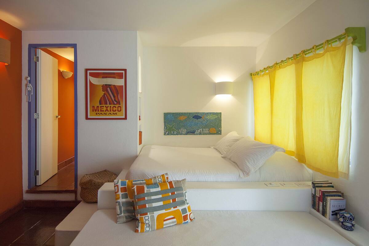 Une chambre à coucher avec un lit, des oreillers, une table de chevet, un tableau, une fenêtre avec des rideaux et une porte.