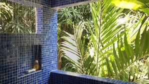 A bathroom with blue mosaic tiles, a shower, and a view of tropical plants.