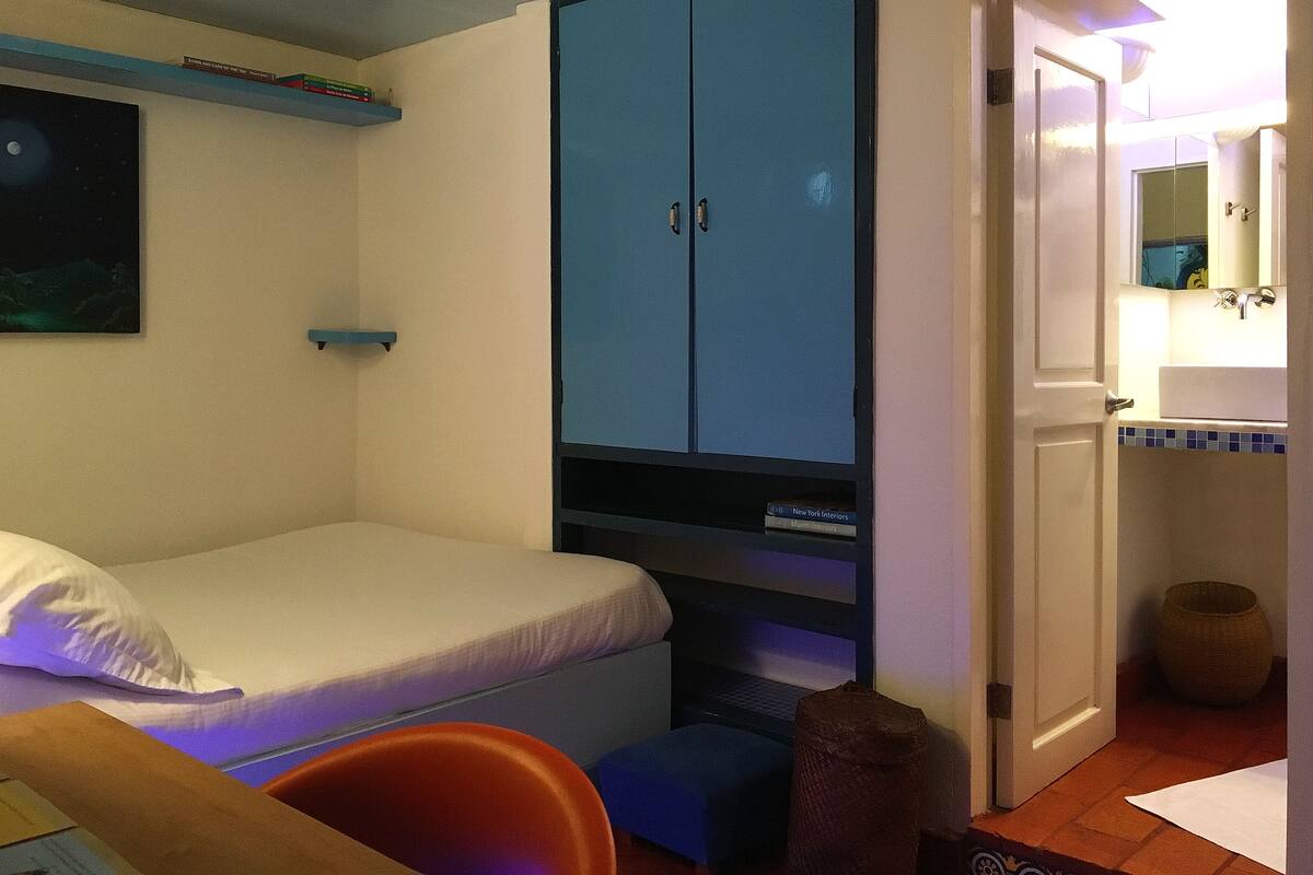 Une chambre à coucher avec un lit, une armoire bleue, un bureau avec une chaise et une salle de bain avec un lavabo et un miroir.