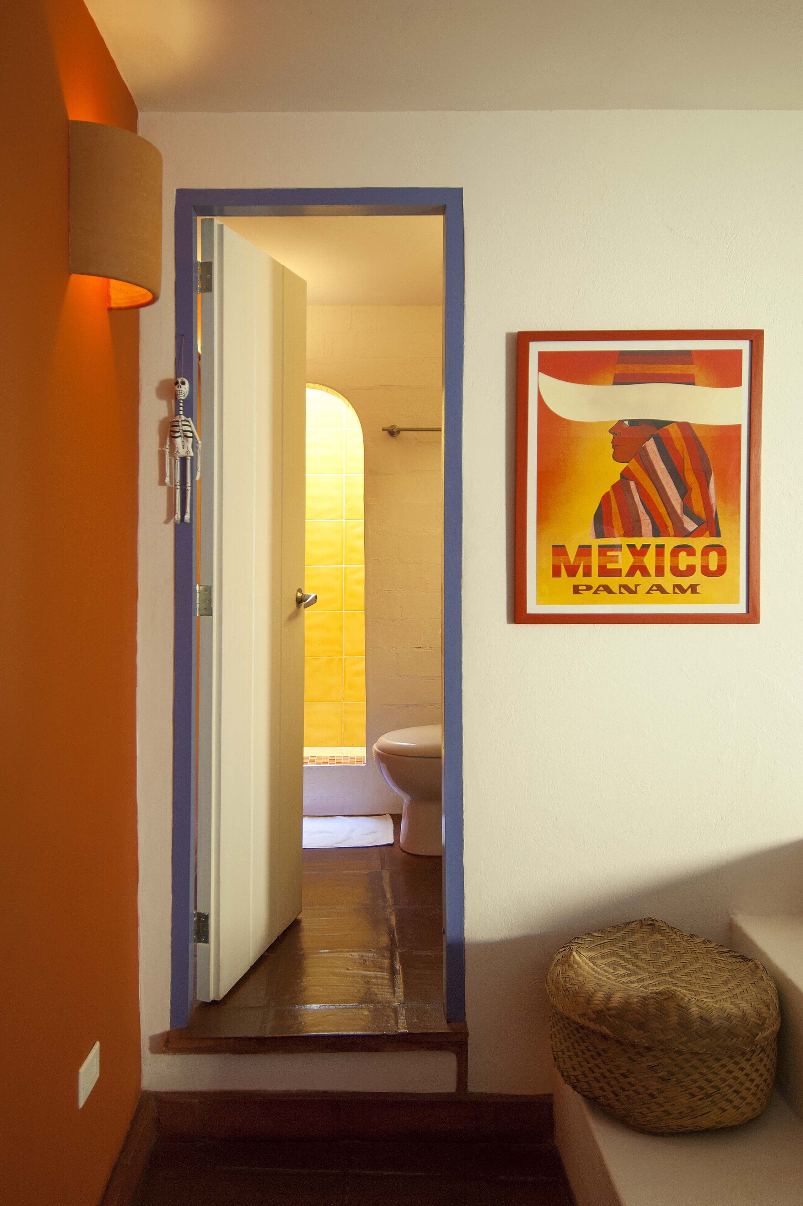 Un couloir avec une porte donnant sur une salle de bain, une affiche encadrée au mur et un panier en osier posé sur le sol.