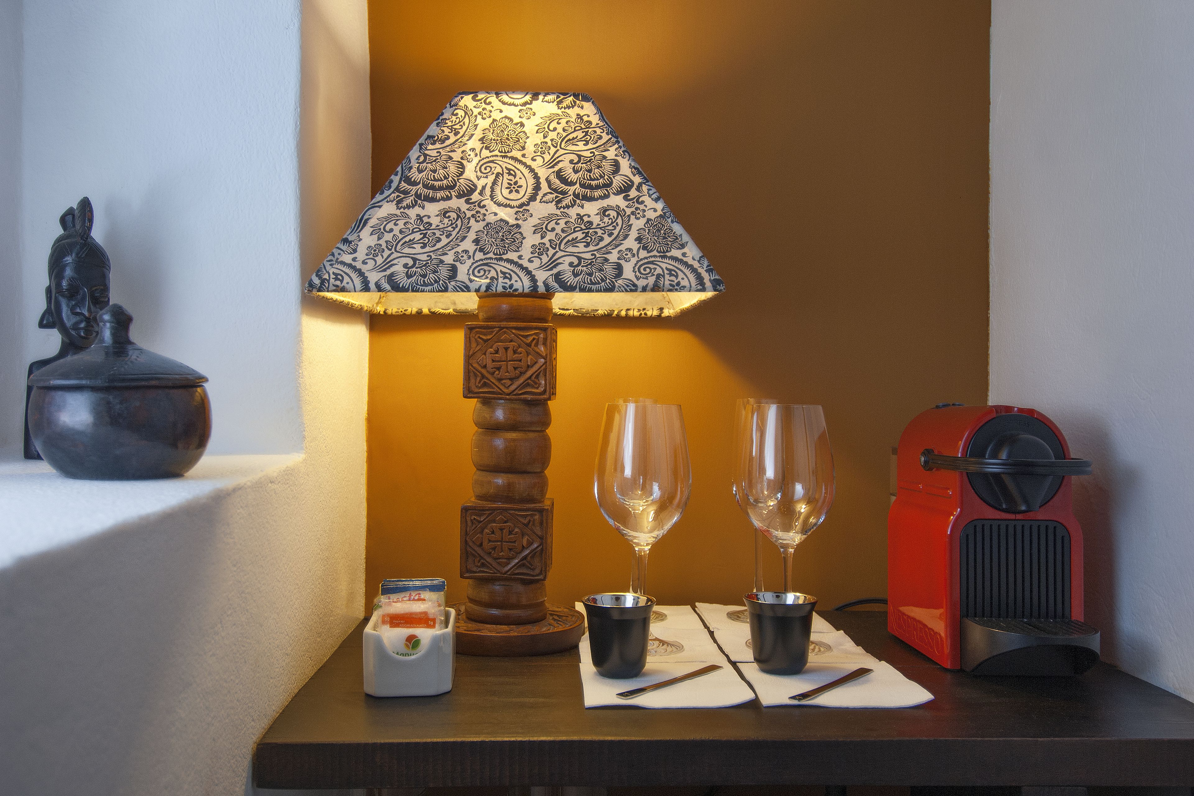 Una mesa con una lámpara, dos copas de vino, una pequeña estatua, un electrodoméstico rojo y un recipiente con una cuchara.