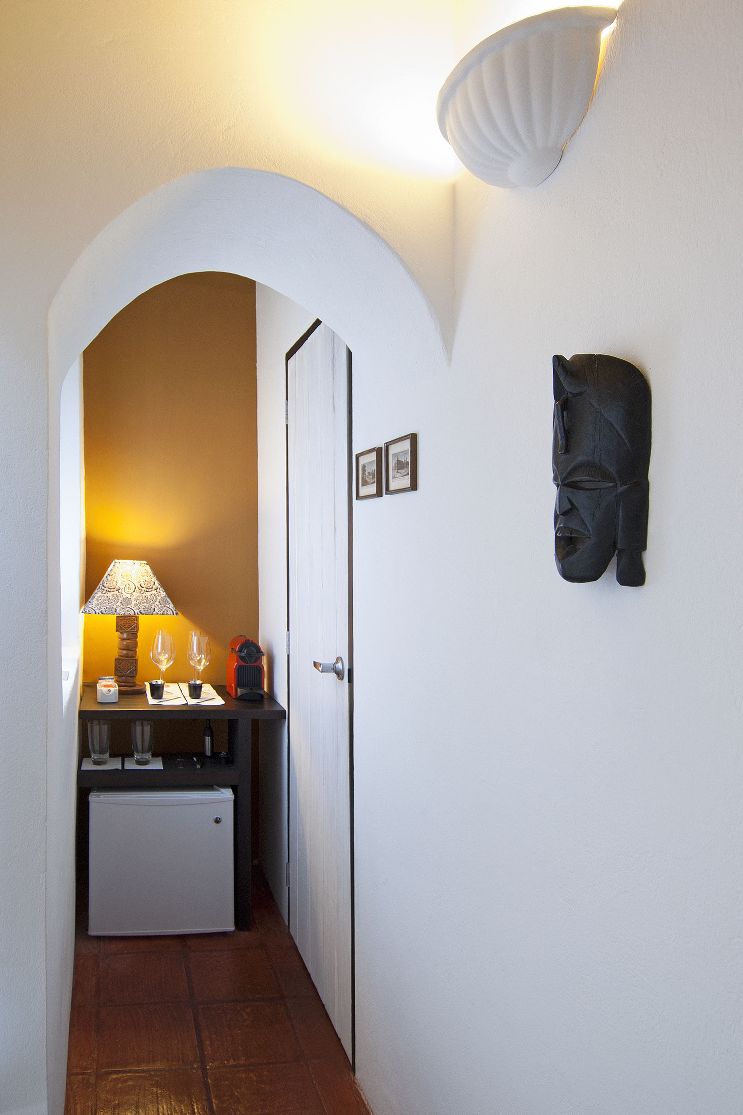 Un pasillo estrecho con una puerta blanca, un refrigerador pequeño, una mesa con lámpara, vasos y una radio antigua.
