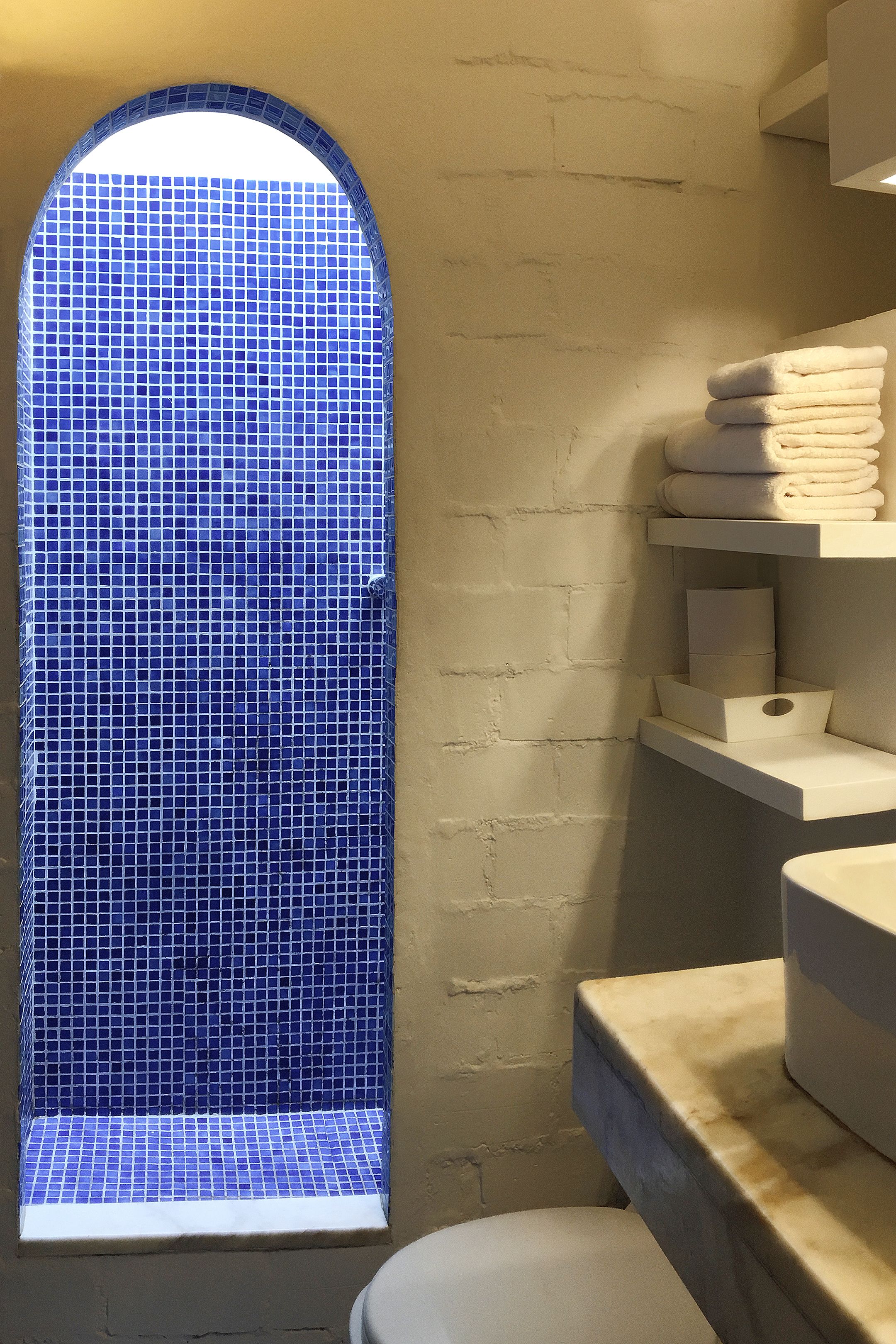 Un baño con pared de azulejos, una ventana y una pila de toallas en un estante.