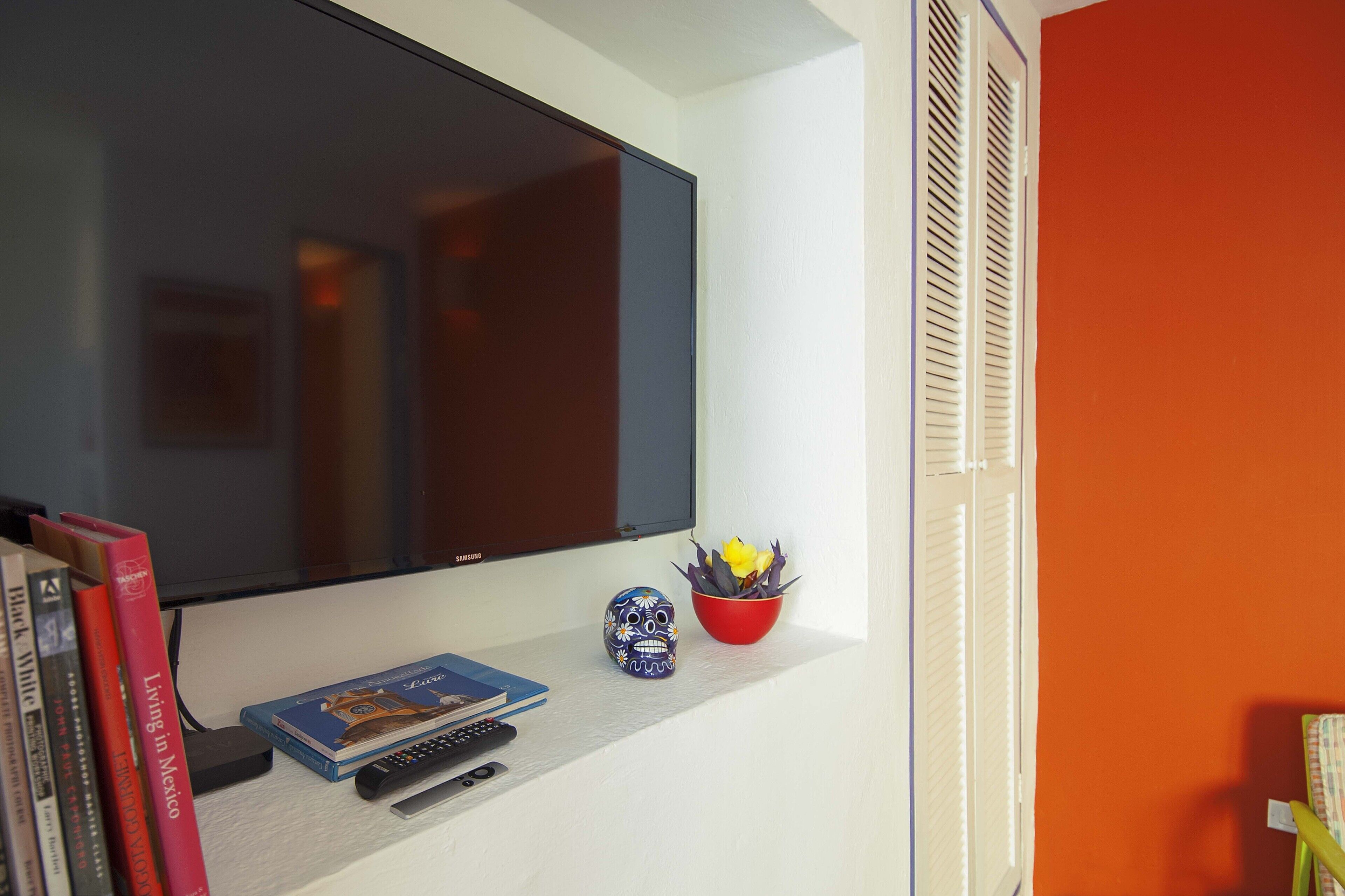Un televisor de pantalla plana montado en la pared, un estante con libros, un control remoto, una calavera decorativa y un cuenco rojo con flores.